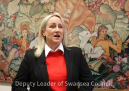 Video screenshot showing Swansea Council Deputy Leader Andrea Lewis in front of a tapestry with the caption Deputy Leader of Swansea Council